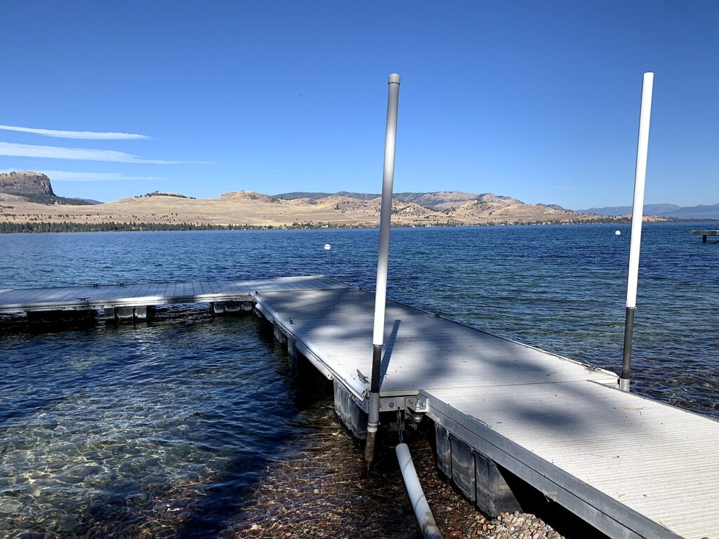A serene lake view showing a wooden dock surrounded by clear water, with hills and a blue sky in the background.