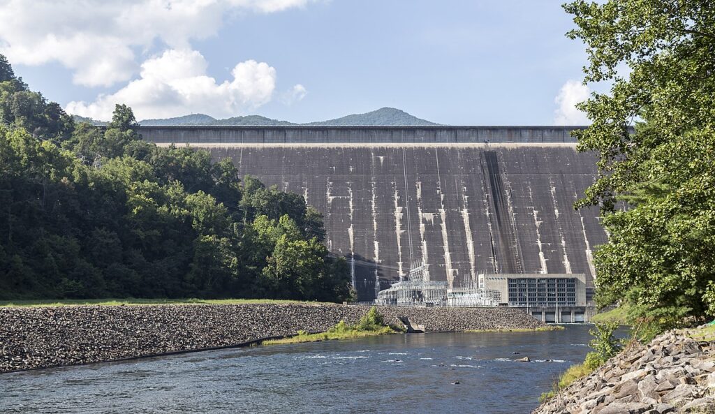 A large dam stands against a backdrop of rolling mountains, with a river and lush trees in the foreground under a blue sky.