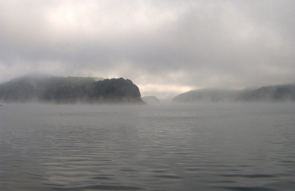 Misty landscape over calm waters, featuring distant, shrouded islands and a blanket of fog enveloping the serene scene.