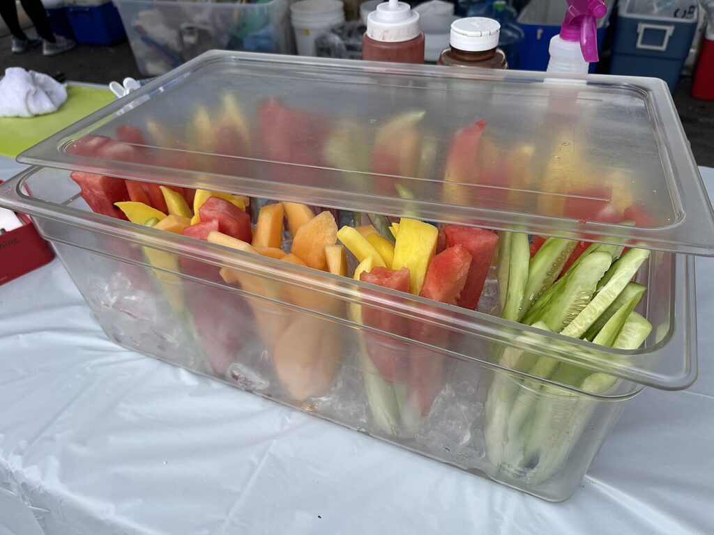 A clear container holds colorful fruit and vegetable sticks, including watermelon, cantaloupe, cucumbers, and yellow melon, on ice.