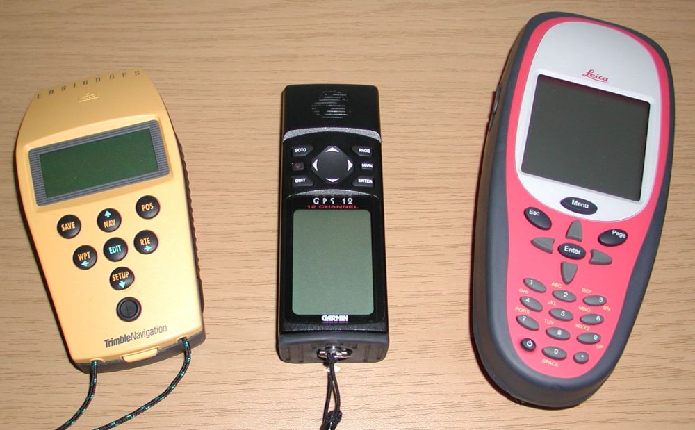 Three handheld GPS devices displayed on a wooden surface: a yellow Trimble, a black Garmin GPS 12, and a red Leica model.