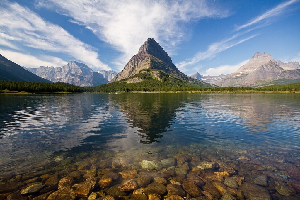 Clear lake reflecting rugged mountains under a bright blue sky, with a foreground of smooth, colorful stones beneath the water.