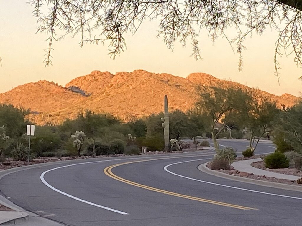A winding road leads through desert vegetation, with sunlit mountains rising in the background under a warm, golden sky.