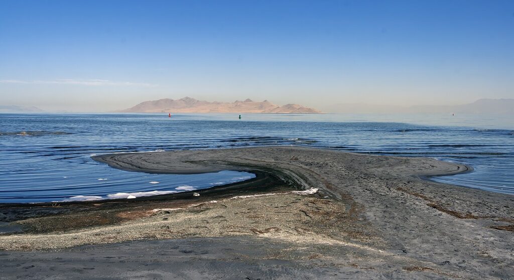 A serene view of a sandy shoreline curving into calm waters, with distant mountains under a clear blue sky.