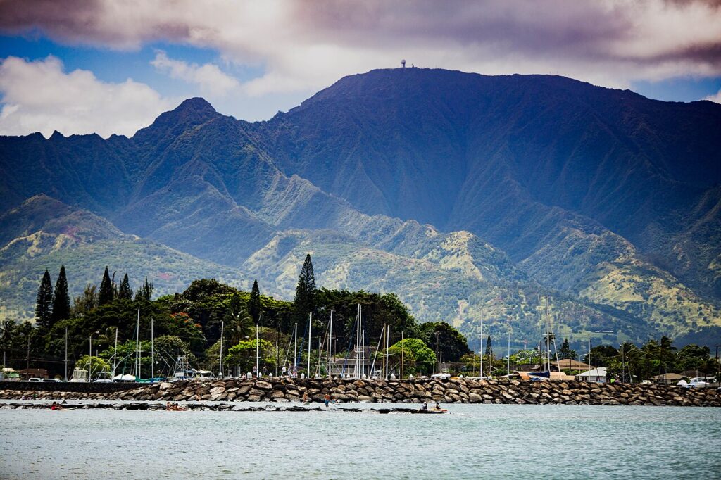 A scenic view of lush mountains towering over a calm harbor filled with sailboats, bordered by a rocky shoreline under a cloudy sky.