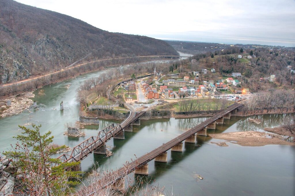 Aerial view of a scenic town by the river, featuring bridges, historic buildings, and surrounding wooded hills under a cloudy sky.