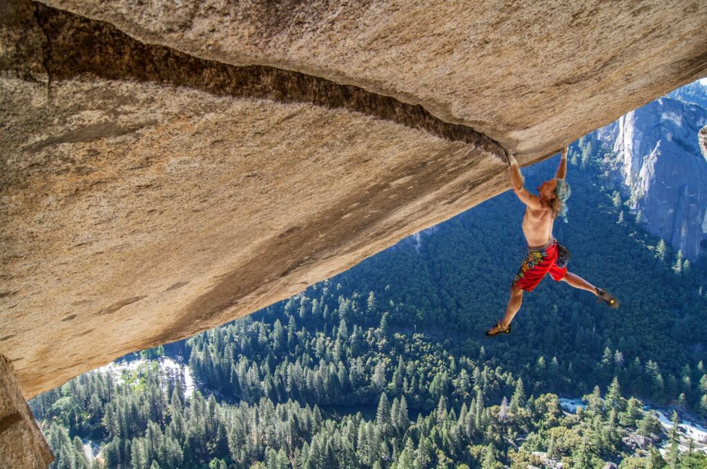 A climber hangs dramatically from a steep rock face in Yosemite, showcasing strength against a backdrop of lush green forests below.