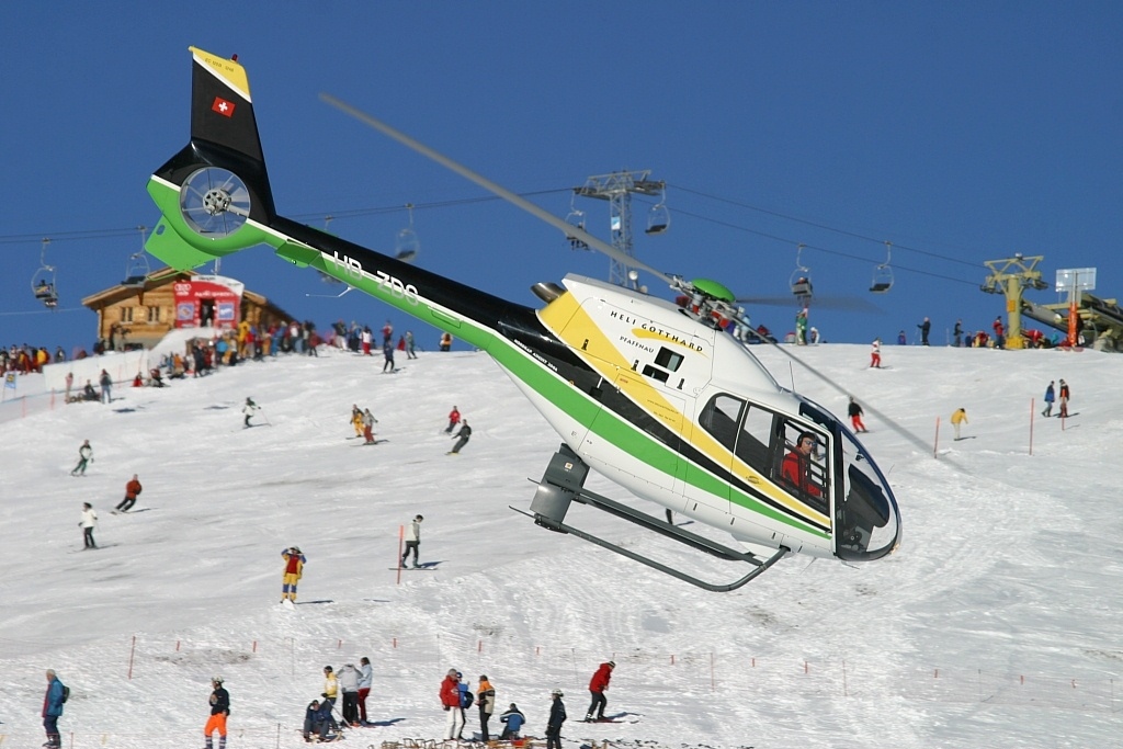 A helicopter hovers above a snowy ski slope, surrounded by skiers and ski lifts under a clear blue sky.