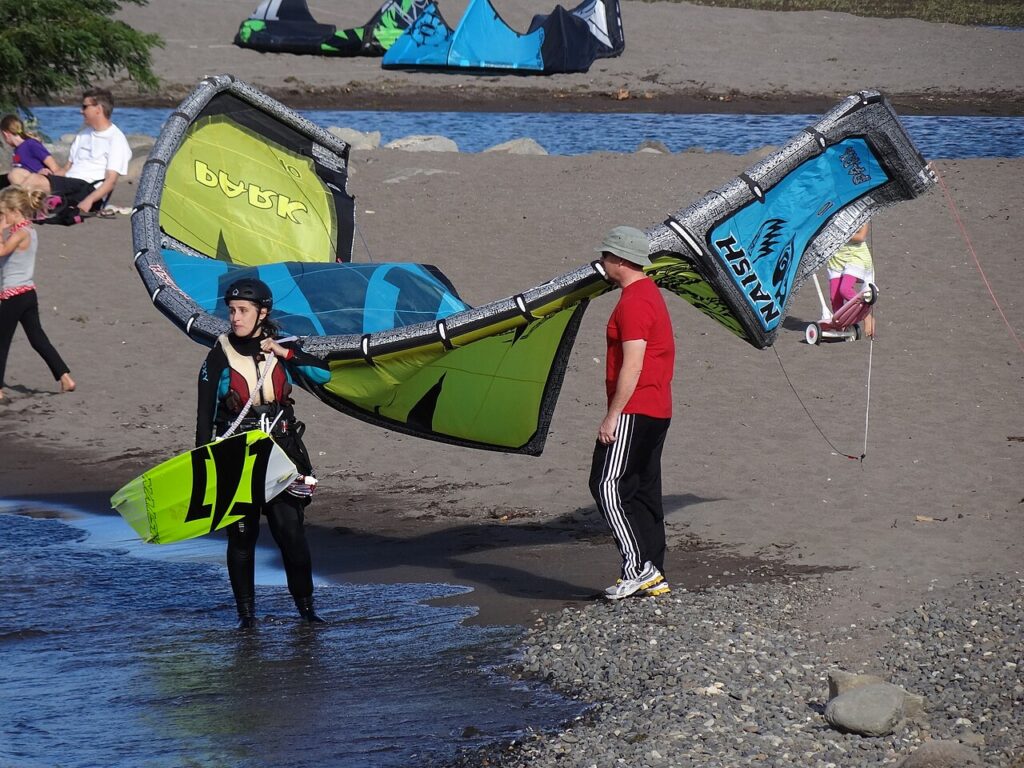 A kitesurfer prepares to launch a vibrant kite on a sandy beach, with spectators nearby and colorful kites in the background.
