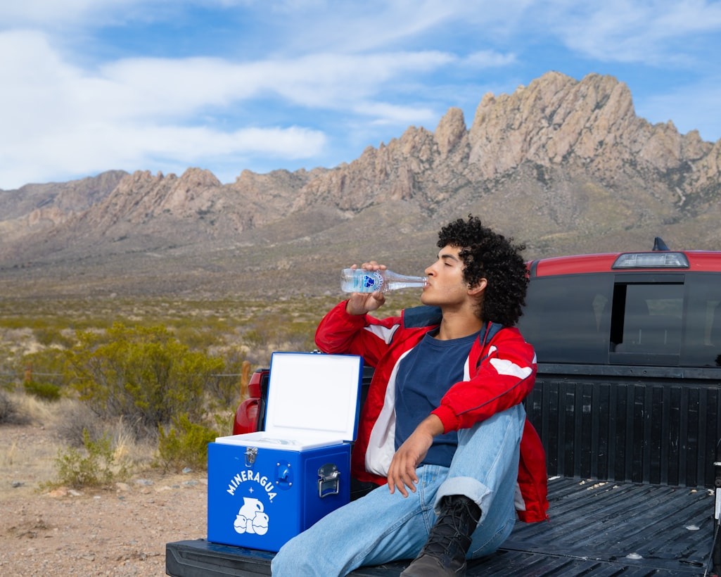 A person sits on a pickup truck's tailgate, drinking water, with a cooler nearby and rugged mountains in the background.