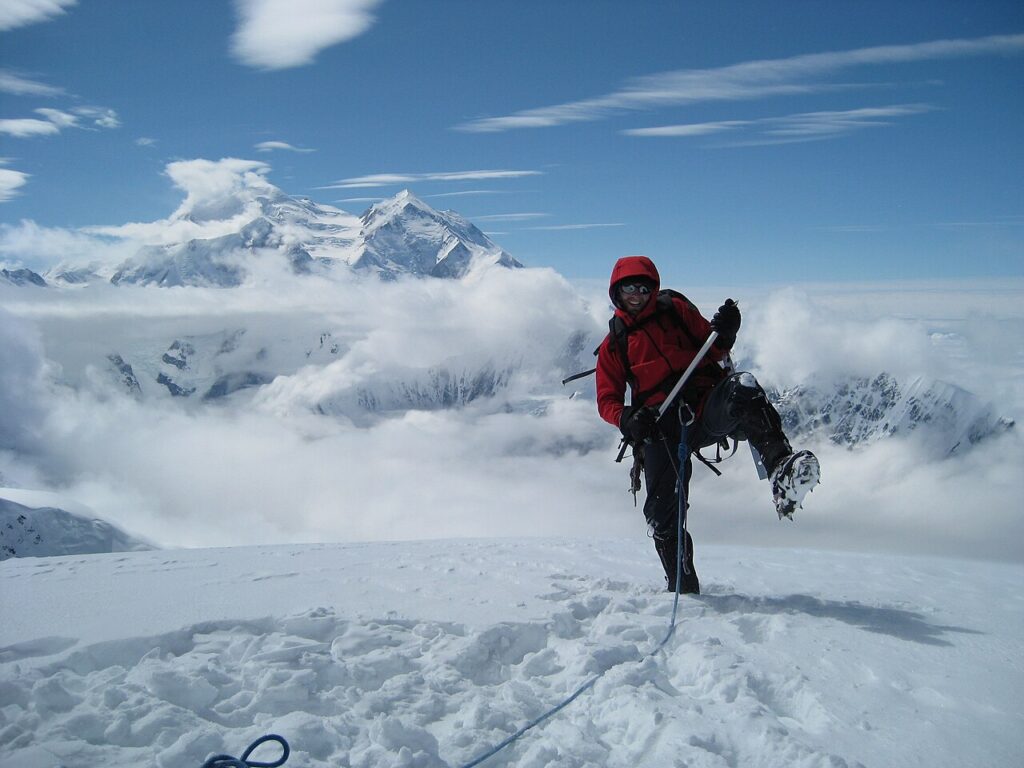 A climber in bright red gear ascends a snowy peak, surrounded by clouds and towering mountains under a clear blue sky.