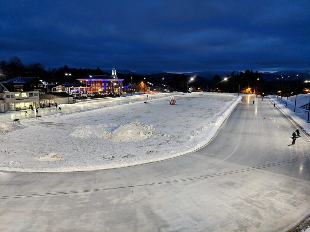 A winter evening skate rink, illuminated by streetlights, surrounded by snow and festive decorations, with skaters gliding on the ice.
