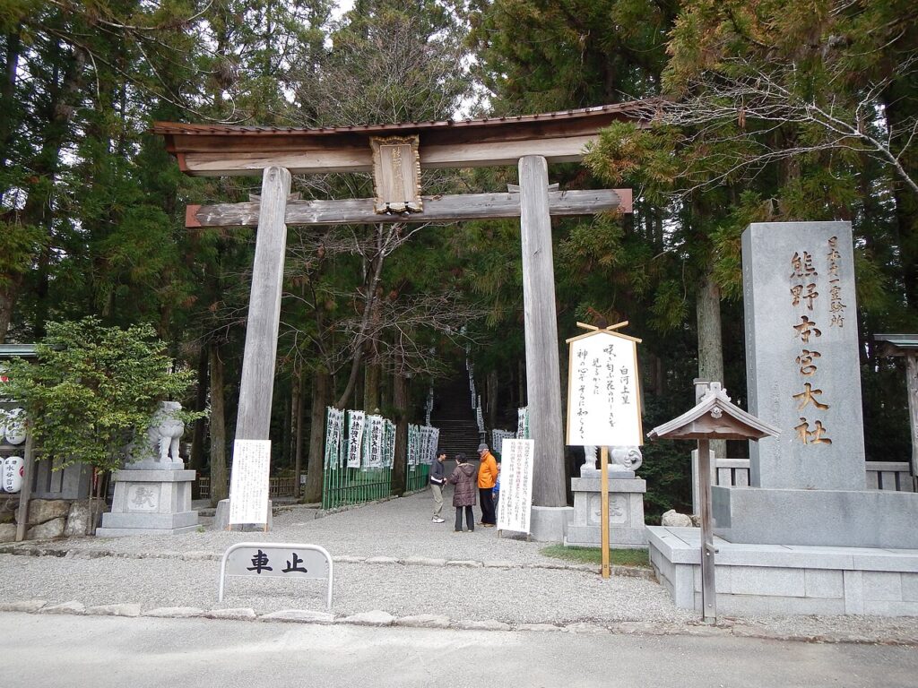 A traditional torii gate marks the entrance to a shrine, surrounded by tall trees, with visitors gathered near stone sculptures.