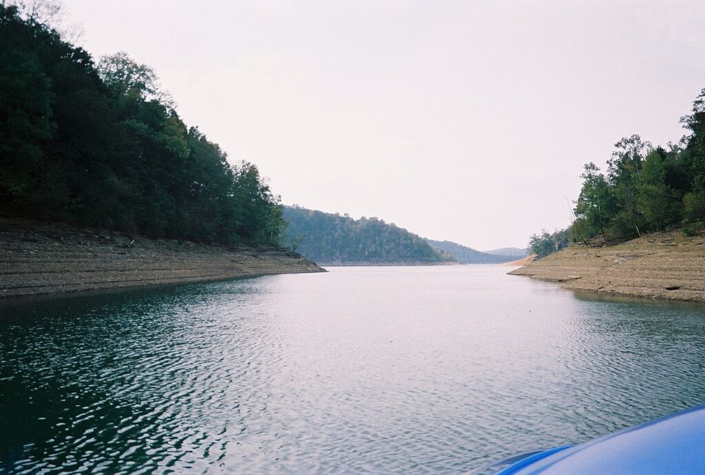 A serene lake view framed by tree-covered hills, with calm waters reflecting the overcast sky and exposed banks along the shoreline.