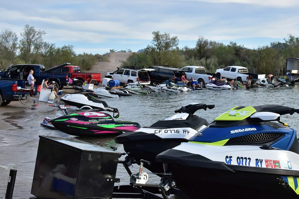 A busy lakeside scene with people loading jet skis onto trucks, surrounded by water and trees under a cloudy sky.