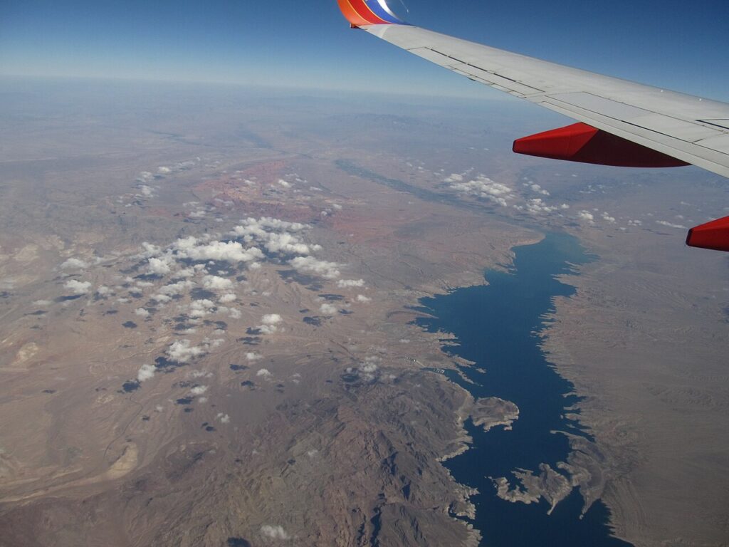 Aerial view of a deep blue reservoir, surrounded by textured desert landscape and scattered clouds, with an airplane wing in the corner.