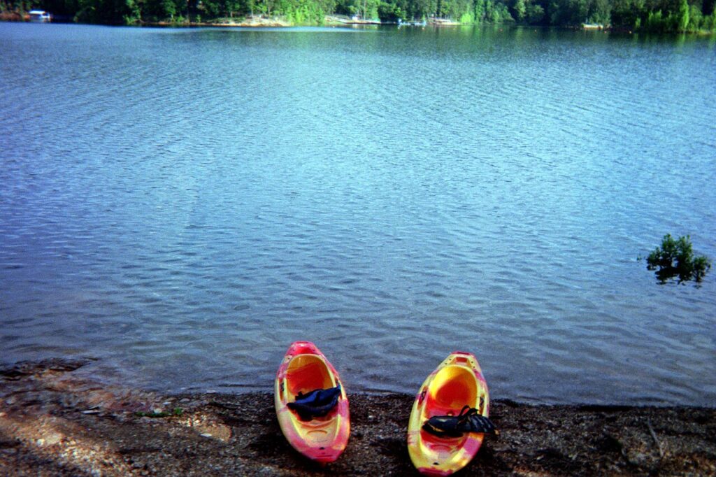 Two colorful kayaks rest on a sandy shore by a calm lake surrounded by lush greenery under a clear blue sky.