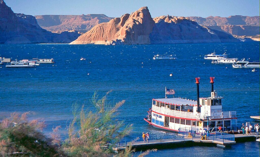 A paddleboat docked at a marina on a lake, surrounded by rocky hills and small boats under a clear blue sky.