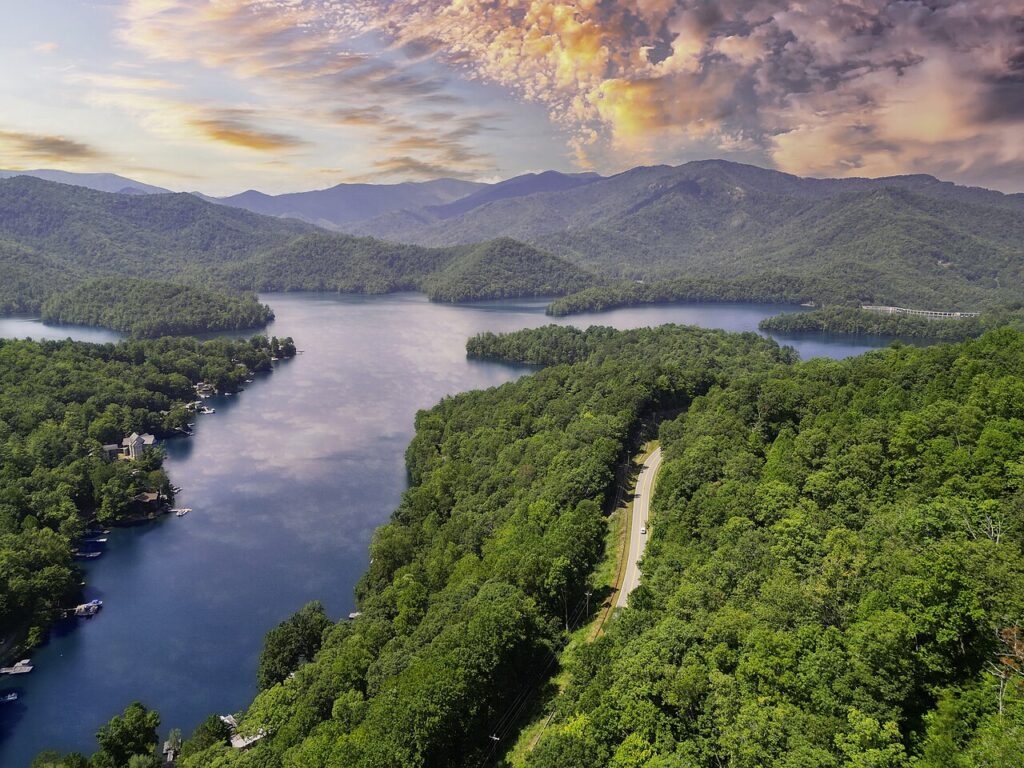 A scenic aerial view of a winding lake surrounded by dense green forests and mountains under a colorful sky at sunset.