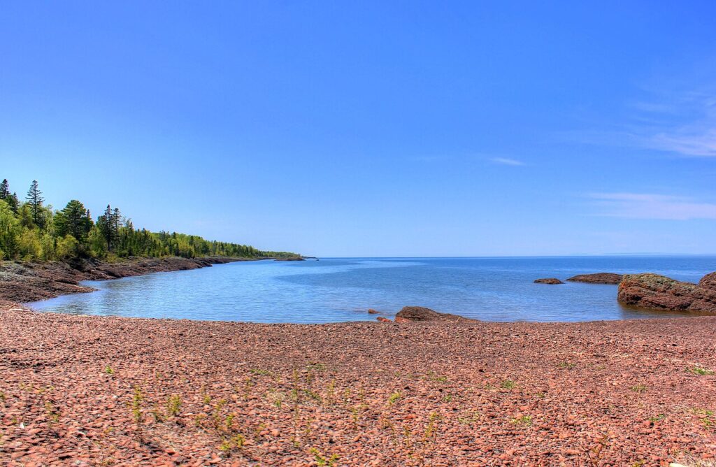 A tranquil shoreline with smooth stones, surrounded by lush green trees under a clear blue sky, meeting calm waters.