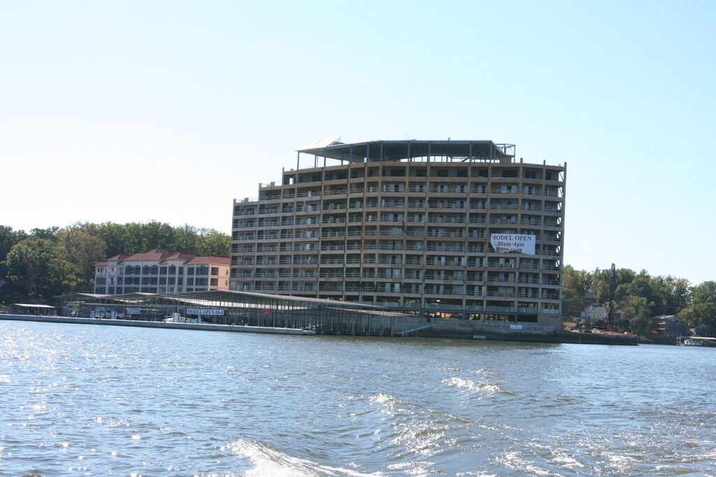 A partially constructed waterfront building with multiple balconies and a "MODEL OPEN" banner, surrounded by trees and water.
