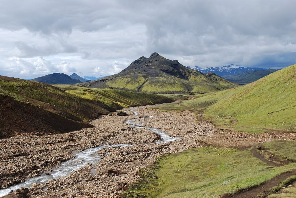 A serene landscape showcasing a winding stream amid green hills and rocky paths, framed by distant mountains under a cloudy sky.