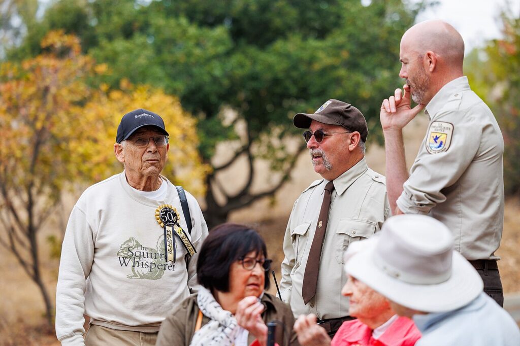 An elderly man in a "Squirrel Whisperer" sweatshirt celebrates with others at an outdoor event, surrounded by autumn foliage.