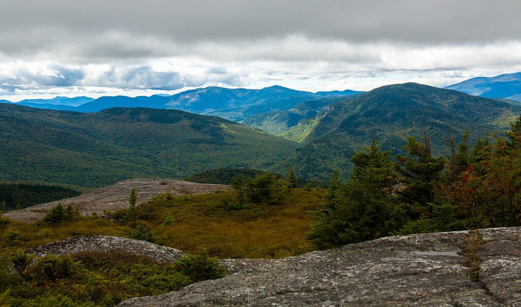Vast mountain landscape under cloudy skies, with rolling green hills and distant peaks stretching into the horizon.