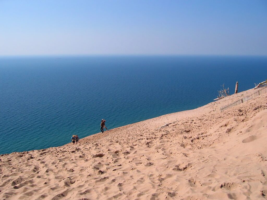A sandy dune slopes toward a serene blue lake, with two hikers making their way up and a clear sky above.