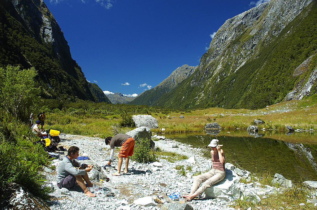 A group of people enjoying a sunny day by a serene lake in a valley surrounded by majestic mountains and lush greenery.