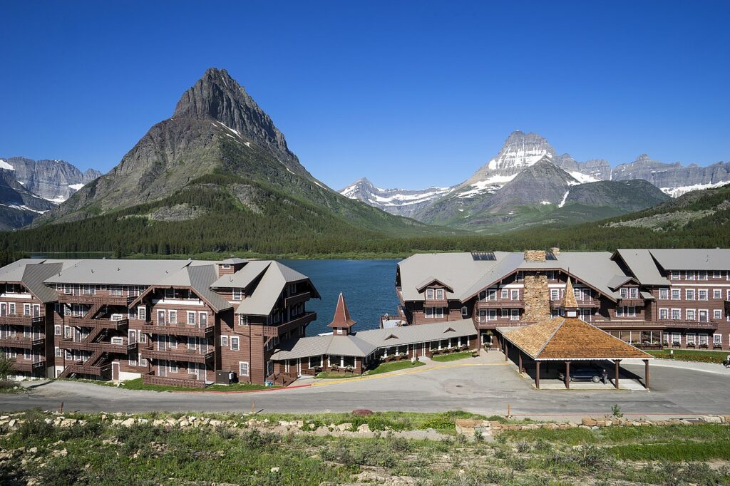 Scenic view of a rustic lodge beside a serene lake, framed by majestic mountains under a clear blue sky.