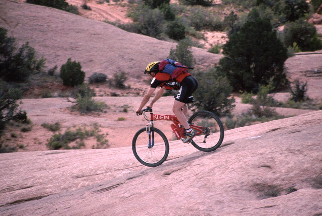 A mountain biker in a red jersey navigates rocky terrain, showcasing skill and determination against a desert landscape.