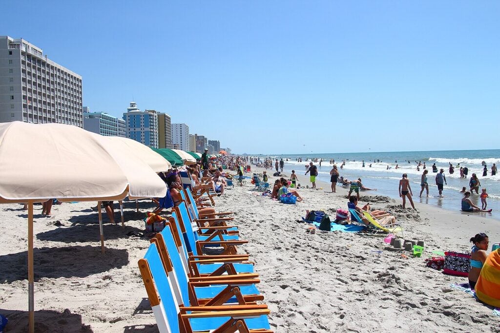 A bustling beach scene with sunbathers, colorful umbrellas, and people enjoying the water, lined with tall buildings under a clear blue sky.