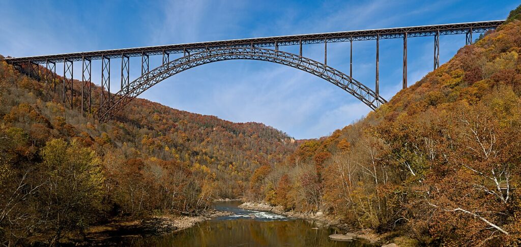 A towering steel arch bridge spans across autumn-colored hills, reflecting in a tranquil river below, under a blue sky.