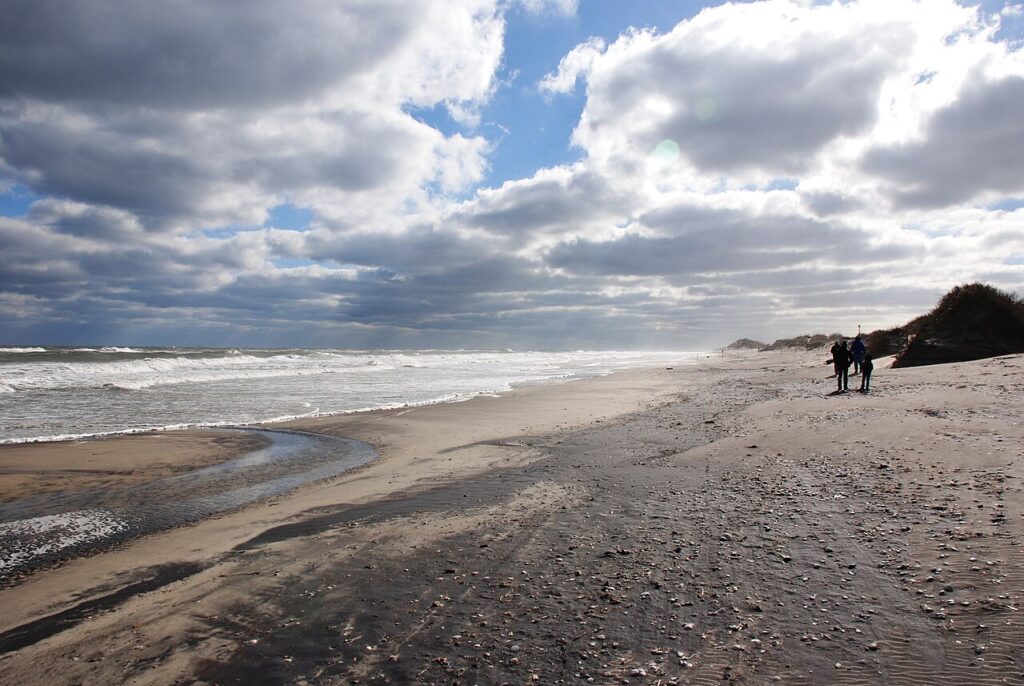 A sandy beach under cloudy skies, with gentle waves lapping at the shore and two people walking in the distance.