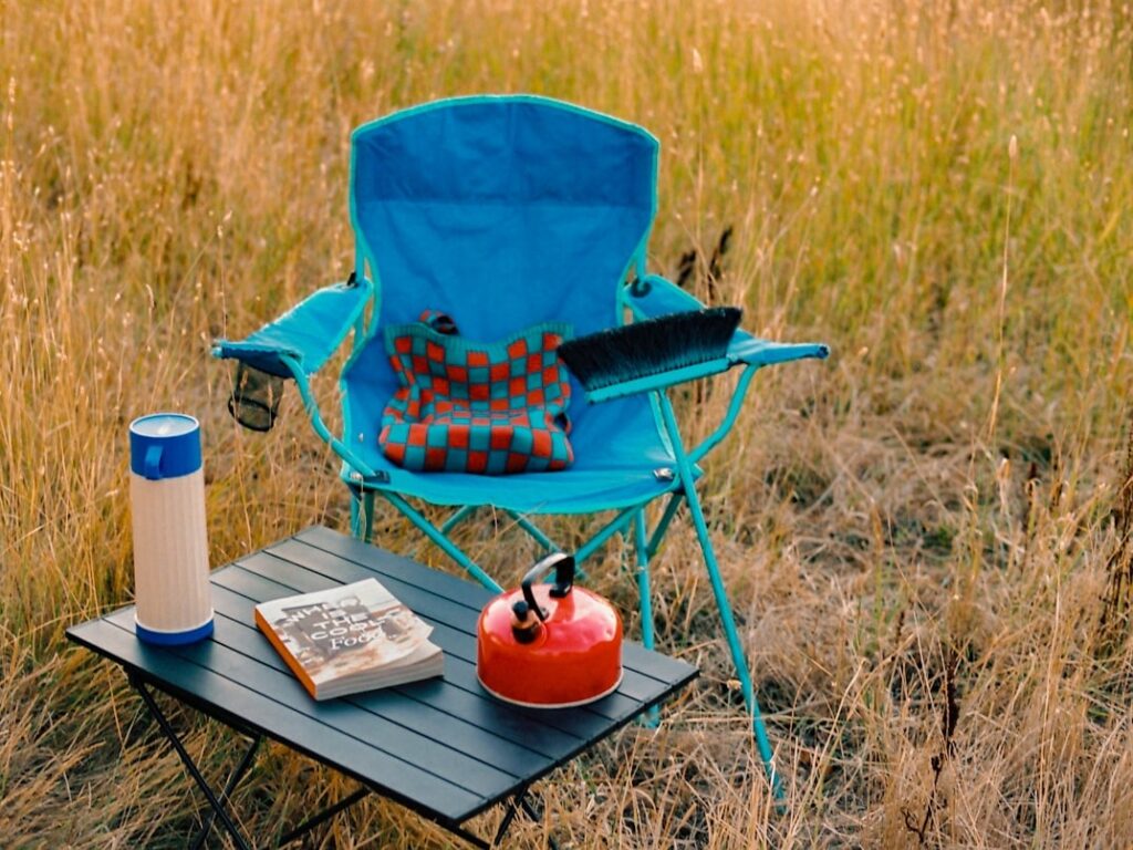 A blue folding chair with a colorful blanket sits beside a small table holding a book, a red kettle, and a lantern in a grassy field.