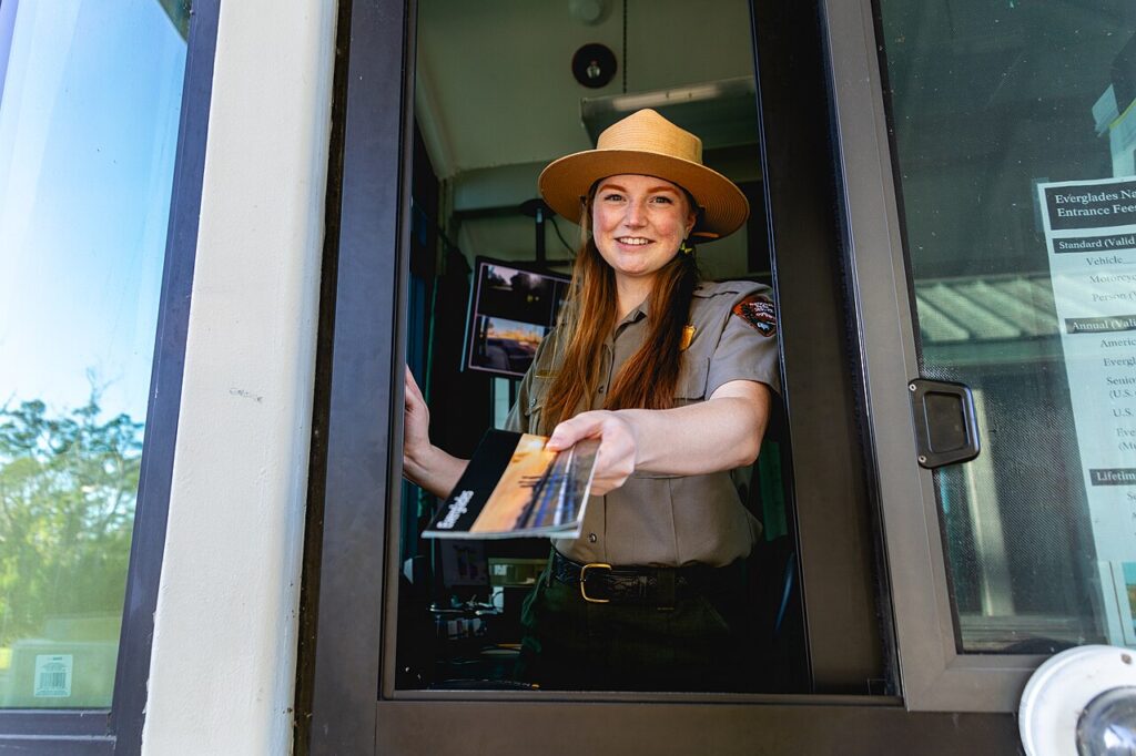 A park ranger in uniform hands out a brochure from a booth window, with park information visible in the background.