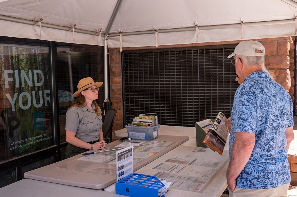 A park ranger sits at a information desk, assisting an older man reviewing park brochures under a tent.