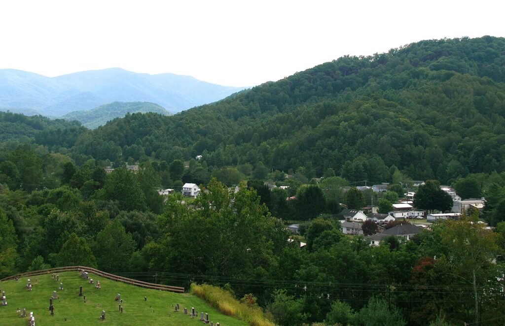 A serene view of a valley surrounded by mountains, featuring a small town and a cemetery on a grassy hill in the foreground.
