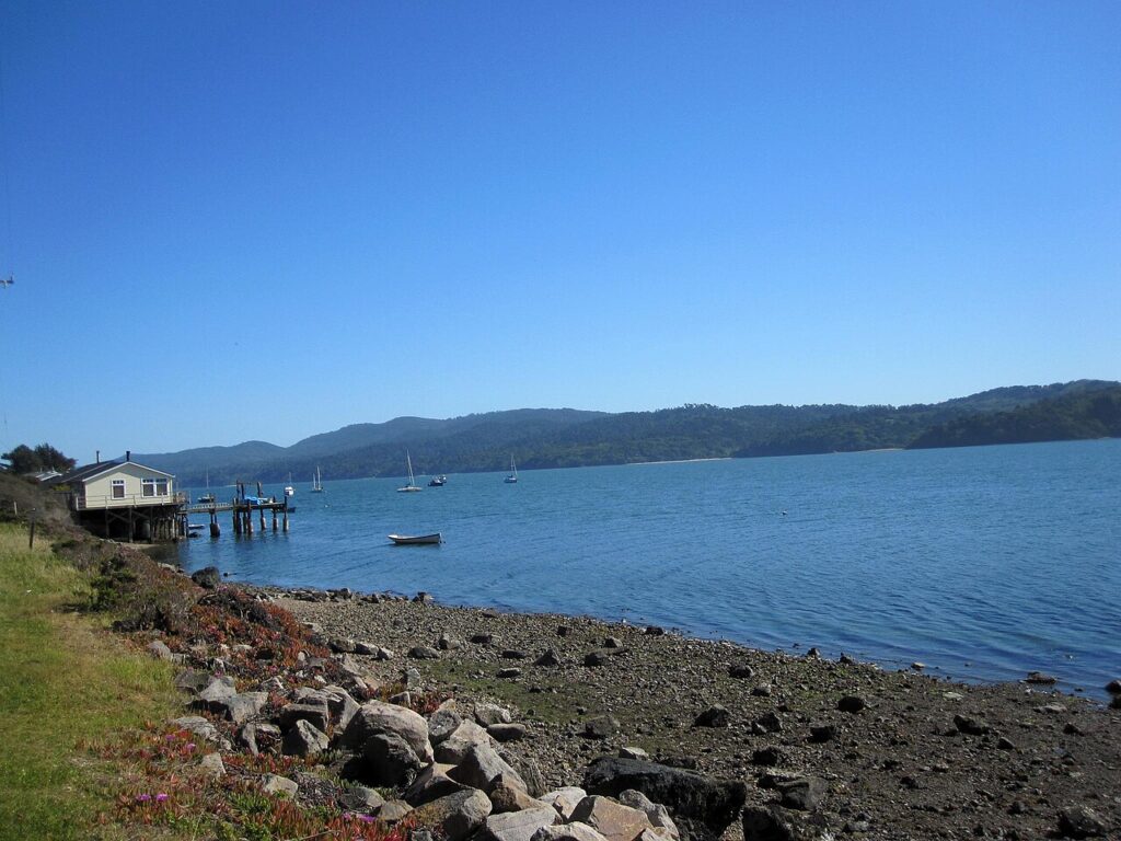 A serene coastal view featuring a small house, boats on calm water, and lush hills under a clear blue sky.