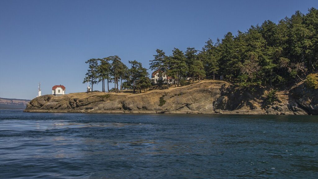 A coastal scene featuring a lighthouse and a white house among trees on a rocky shoreline, under a clear blue sky.