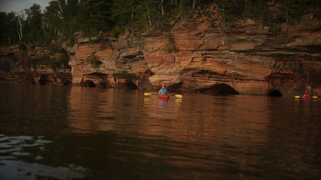 Two kayakers paddle in tranquil waters near stunning sandstone cliffs, surrounded by lush greenery on a calm day.