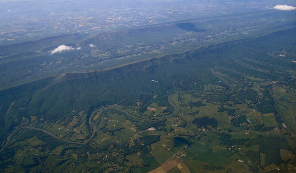 Aerial view of a winding river flowing through lush green farmland and rolling hills, with distant mountains under a clear blue sky.