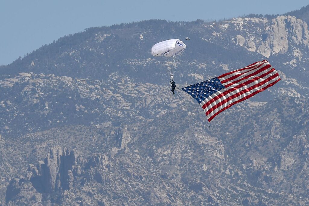 A skydiver descends with a large American flag attached to their parachute against a backdrop of rugged mountains.