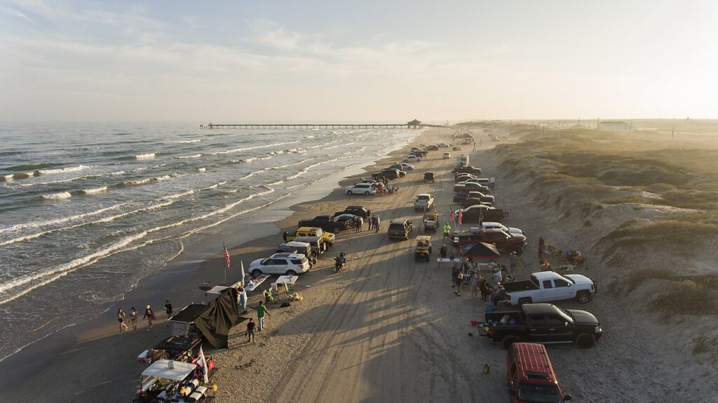 Aerial view of a lively beach scene with parked cars, people walking, and a distant pier under a warm sunset sky.