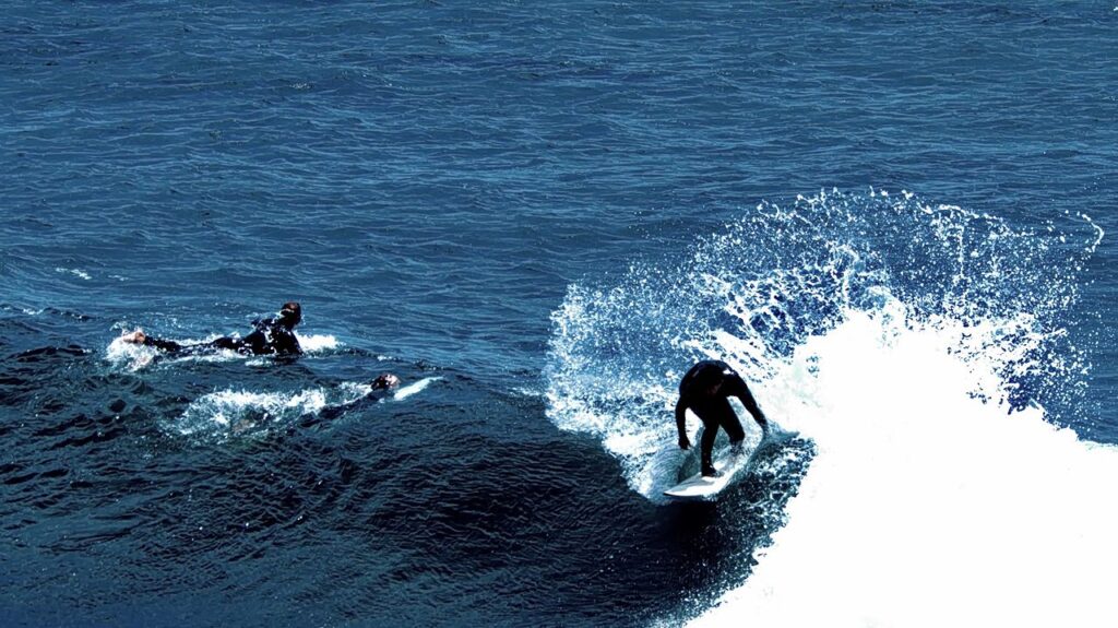 A surfer in a black wetsuit rides a wave while two other surfers swim nearby in the deep blue ocean.