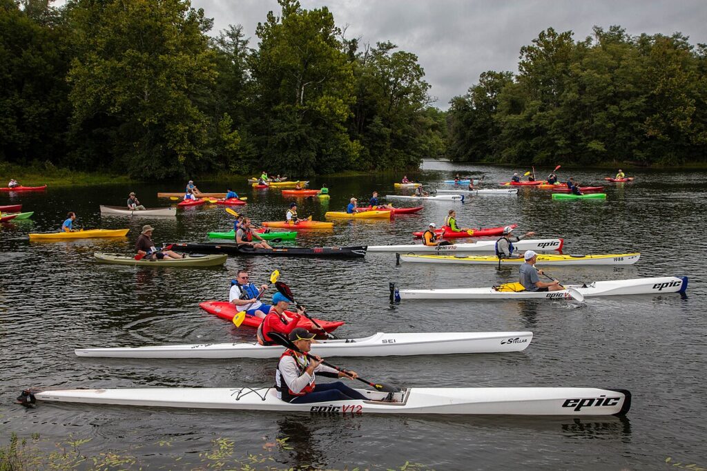 A vibrant group of kayakers in various colored boats paddling on a calm river surrounded by lush green trees under a cloudy sky.