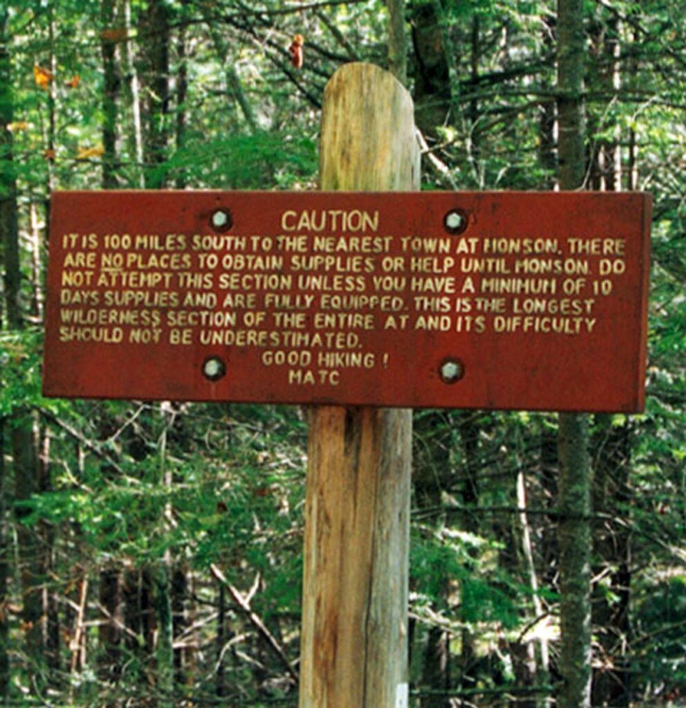 A weathered red sign warns hikers about a 100-mile stretch without supplies, urging proper preparation for the challenging section ahead.