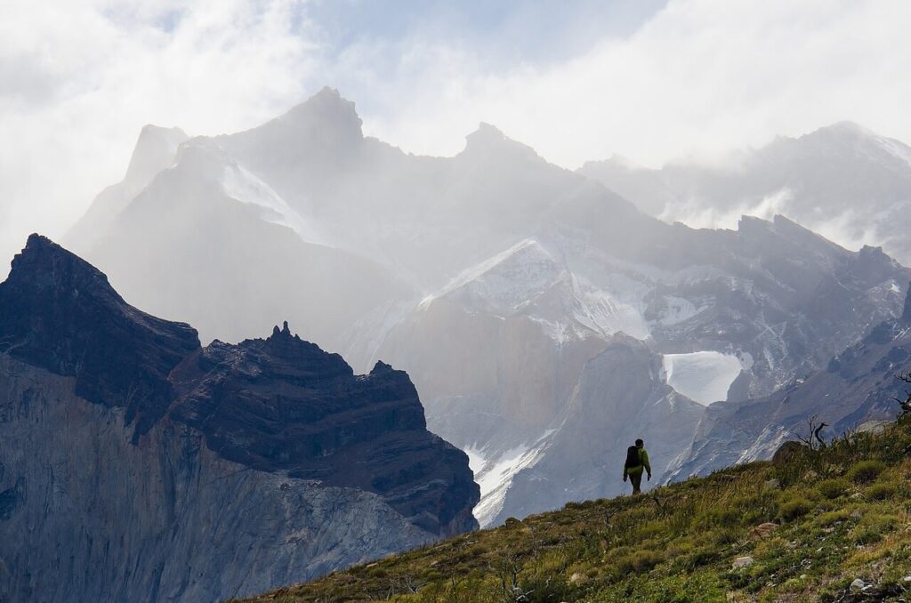 A hiker in a green jacket traverses a rugged mountain landscape shrouded in mist and towering peaks under a cloudy sky.