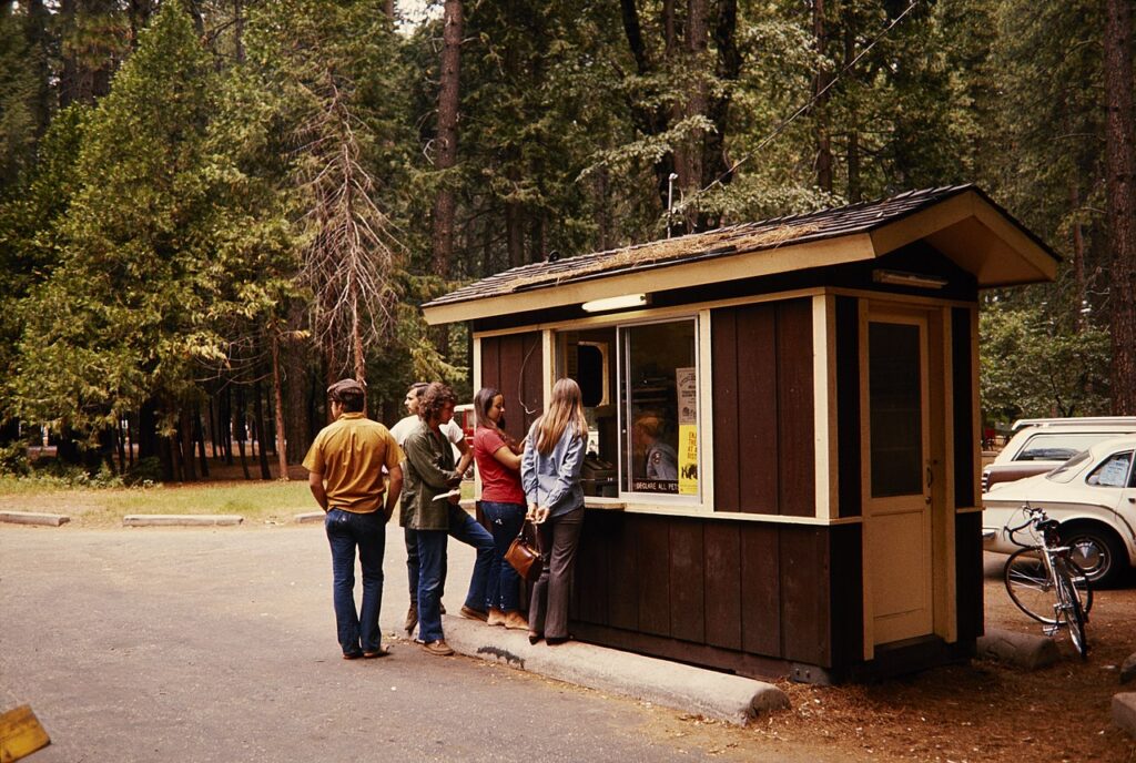 A group of four people waits at a wooden kiosk in a forested area, with cars and bicycles in the background under towering trees.